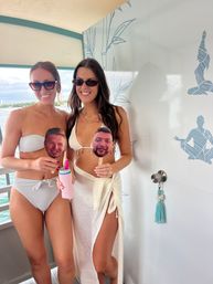 Two smiling women in white swimwear and sunglasses pose on a boat deck by the water, holding novelty face-on-a-stick props and a pink insulated tumbler, with blue decorative wall art in the background.