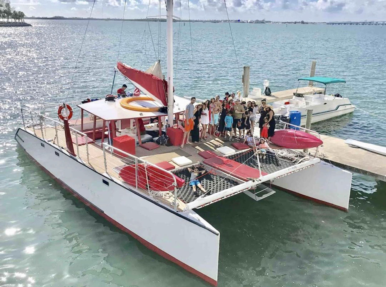 White-and-red catamaran docked at a coastal marina in turquoise water, a group of people posing and lounging on deck nets on a sunny day