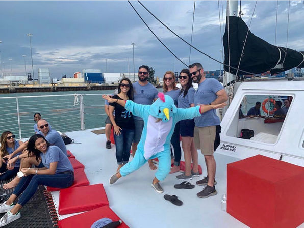 Group of friends on a sailboat deck in a harbor, laughing and posing while someone in a bright blue unicorn onesie strikes a playful pose under a cloudy sky.