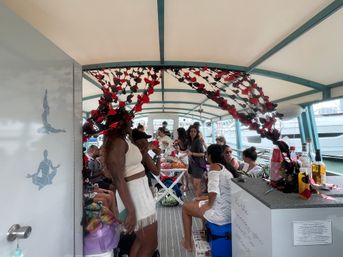 Lively daytime boat party on a covered passenger vessel at a marina, people in summer outfits chatting and enjoying drinks and snacks beneath red-and-black hanging ribbon decorations.