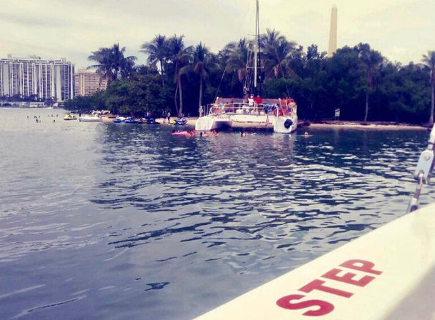 Catamaran anchored off a small palm-lined island with swimmers and jet skis, city high-rises on the distant shoreline, view from a nearby boat rail labeled STEP.