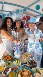 Three women on a boat deck toasting with champagne flutes beneath a "Birthday" banner, wearing summer hats at a coastal yacht birthday party with a colorful snack buffet and life preserver