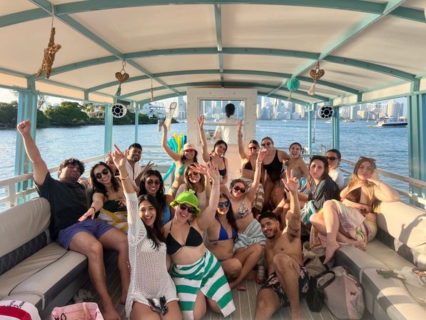 Sun-soaked boat party: a large group of people in swimwear on a covered leisure boat with a coastal city skyline across the bay, waving and smiling.
