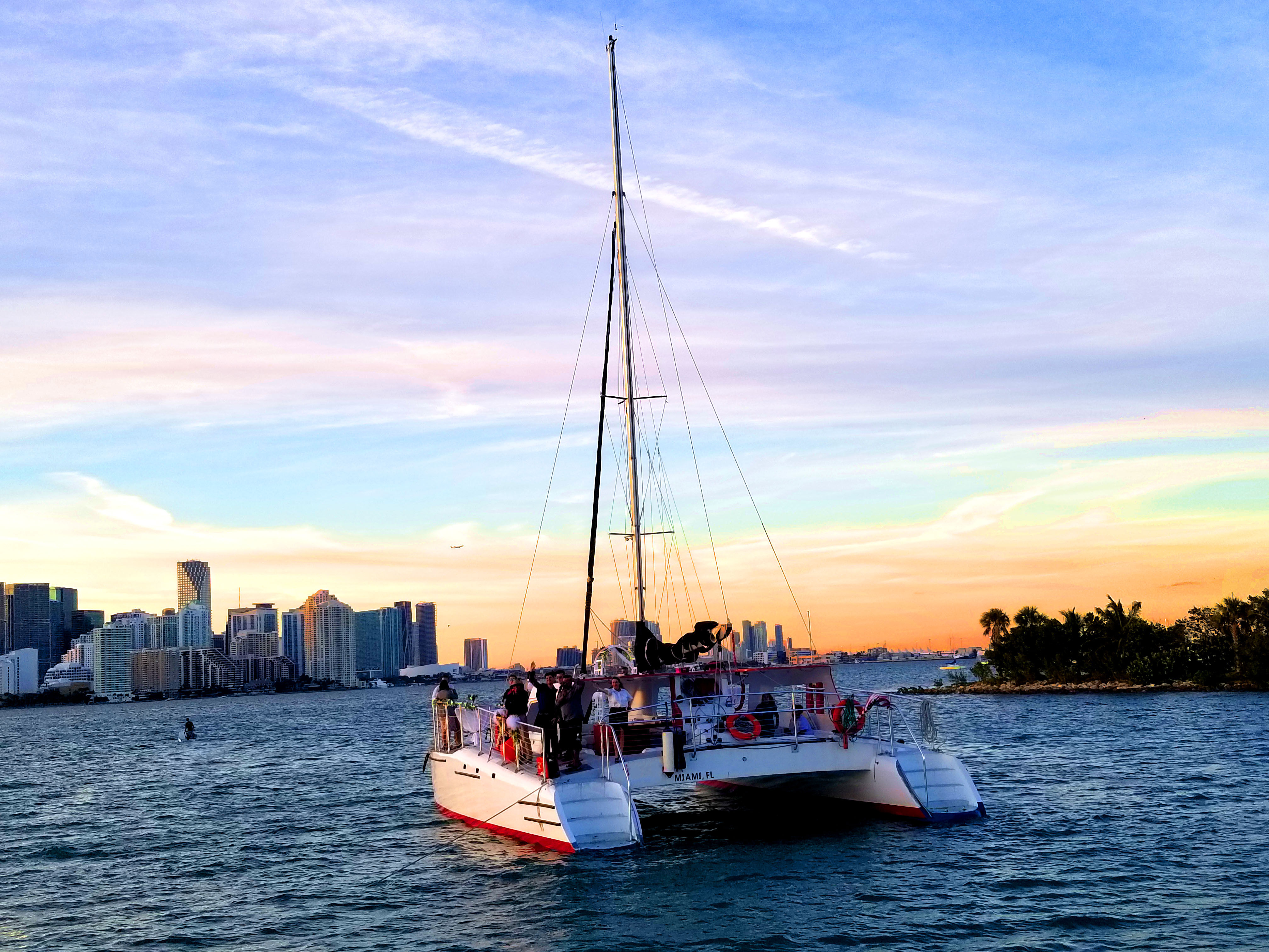 Catamaran sailing in a calm bay at sunset with the Miami skyline and palm‑fringed shore, colorful pastel sky and passengers on deck.