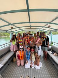 Smiling group of friends on a covered pontoon boat in turquoise tropical waters, wearing colorful Hawaiian leis and novelty sunglasses for a lively summer boat party near a green shoreline.