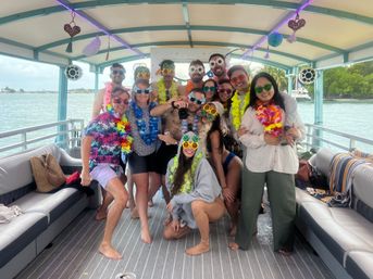 Group of partygoers in colorful leis and novelty sunglasses posing on a decorated pontoon boat during a tropical nearshore cruise with palm-lined shoreline and turquoise water