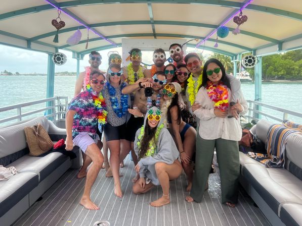Group of partygoers in colorful leis and novelty sunglasses posing on a decorated pontoon boat during a tropical nearshore cruise with palm-lined shoreline and turquoise water