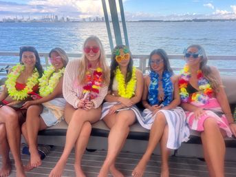 Six friends wearing colorful leis and playful sunglasses sitting on a boat with towels on their laps, smiling as a harbor and city skyline stretch across the sunny bay