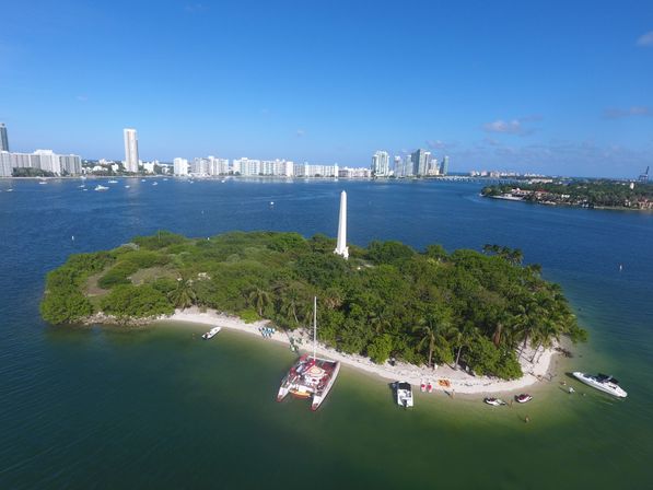 Drone view of a small tropical island with a white obelisk, palm trees and sandy beach, anchored catamaran and boats in a blue bay with a distant coastal city skyline under a clear sky