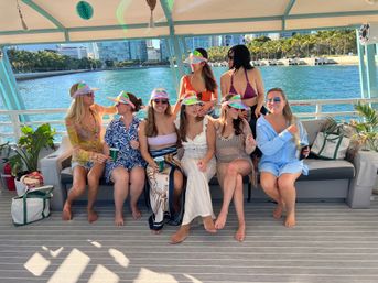 Eight women in colorful sun visors and summer outfits smiling on a covered boat lounge, holding drinks with a sunny waterfront city skyline and palm trees in the background.