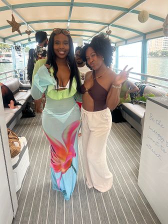 Two friends posing on a covered boat party at a marina — one in a turquoise floral maxi dress and the other in a brown cutout top with white pants, city waterfront visible through windows and festive decorations overhead.