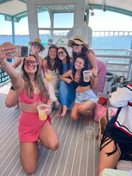 Group of six women taking a selfie on a sunny boat deck, holding drinks and wearing summer outfits, sunglasses and straw hats, with blue water and a distant bridge in the background — relaxed coastal summer vibe.