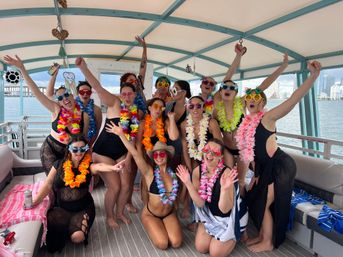 Group of women on a covered party boat wearing swimsuits, bright Hawaiian leis and fun sunglasses, cheering and posing for a bachelorette/girls-trip photo with a coastal city skyline and water in the background.