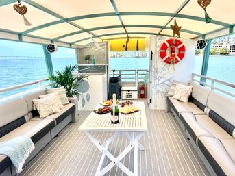 Sunny pontoon boat lounge on a blue bay with a coastal skyline and bridge in the distance — cushioned wraparound sofas, decorative pillows, potted palm, life ring, and central table with champagne and appetizers.