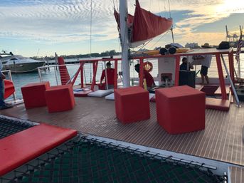 Sunset catamaran deck at a marina with red cube seats and cushions, netted foredeck, furled red sail and yachts on calm water