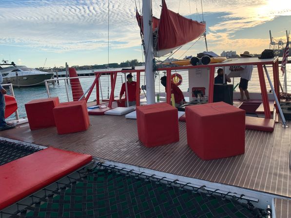 Sunset catamaran deck at a marina with red cube seats and cushions, netted foredeck, furled red sail and yachts on calm water