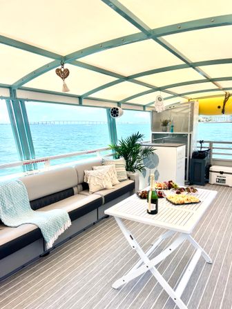 Covered pontoon boat lounge with teal frame and cushioned sofa, cozy throws and pillows, white folding table set with champagne and cheese platter, potted plant, and turquoise bay with a distant bridge on a sunny day.