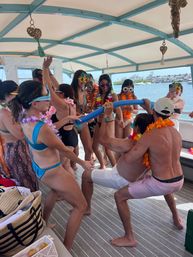 Boat party scene: people in swimsuits wearing colorful leis and novelty sunglasses playing limbo with a pool noodle on a covered party boat near a sunny coastline and marina