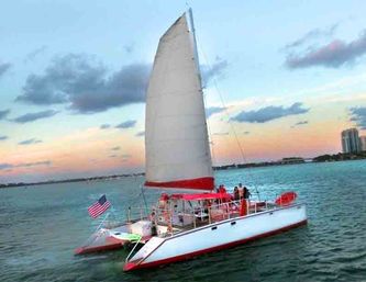 White-and-red catamaran sailboat with tall sail and American flag gliding on turquoise coastal waters at sunset with people on deck and distant skyline