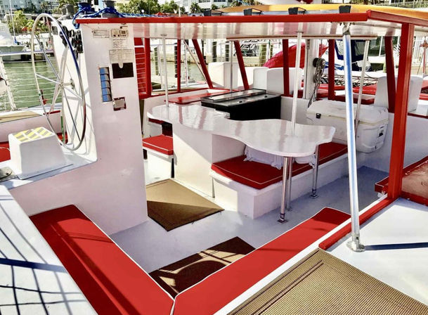 Sunny marina boat deck with red-and-white cushioned benches, curved white dining table, helm wheel and stainless support poles, docked boats visible in the background.