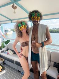 Smiling pair wearing pineapple novelty sunglasses at a sunny boat party — leopard-print bikini, swim trunks and a light robe, marina and water in the background