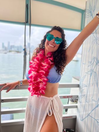 Smiling woman in a blue bikini top and white sarong wearing teal heart-shaped sunglasses and a bright pink lei, posing on a boat deck with turquoise bay water and a coastal city skyline in the background.