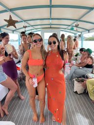Two smiling women in bright orange swimwear and a crochet cover-up holding drinks aboard a covered party boat, friends chatting in the background and water visible beyond the railing.