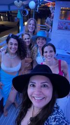 Six smiling friends taking a selfie on a decorated boat at a marina at night with blue lighting and water reflections