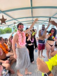Adults on a covered party boat raising champagne glasses, wearing colorful leis and novelty sunglasses, tropical decor with waterfront and shoreline in the background.