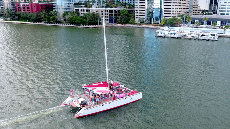 Pink-accented catamaran with passengers cruising a sunny downtown waterfront bay past high-rise buildings and a marina