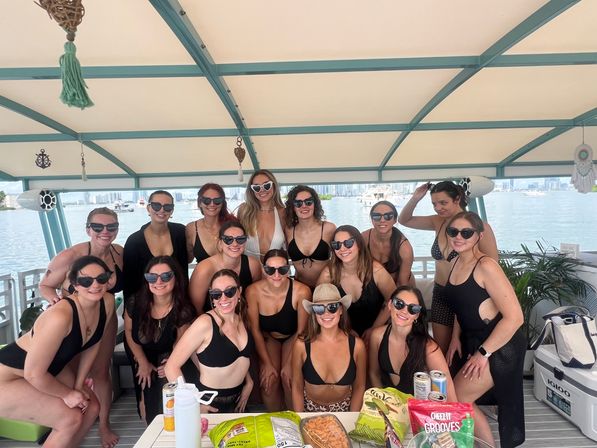 Large group of women in black swimsuits and sunglasses posing and smiling on a covered party boat at a waterfront with a city skyline in the background and snacks and drinks on a table in the foreground.