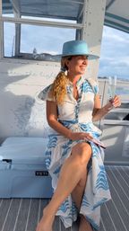 Smiling woman in a blue wide-brim hat and blue-and-white sundress sitting on a sunlit boat deck, holding a champagne flute with ocean and sky in the background.