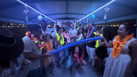 Group of people in colorful Hawaiian leis doing limbo under a blue pool noodle on a lit-up party boat at night with city lights on the waterfront