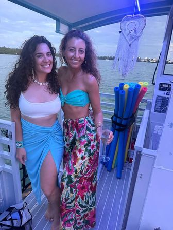 Two smiling women in summer outfits on a boat deck over calm water at dusk — one in a turquoise bandeau and floral skirt holding a champagne flute, the other in a white crop top and blue wrap skirt; colorful pool noodles and a macramé heart hanging nearby.