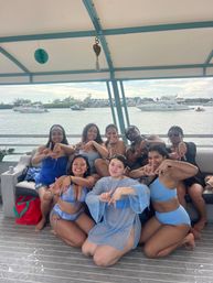 Group of eight friends in blue swimsuits smiling and making hand signs while seated on a covered party boat deck, with yachts and a calm coastal bay visible in the background on a sunny day.