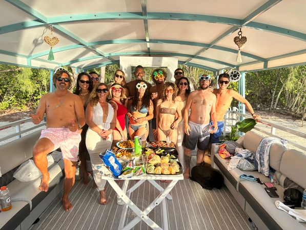 Sunny tropical pontoon boat party with a group of friends in swimsuits and sunglasses posing around a table of snacks and sandwiches, covered deck, sandy shoreline and mangrove trees visible in the background.