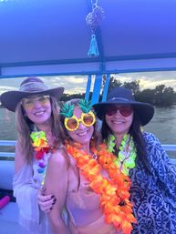 Three friends in wide-brim hats, pineapple sunglasses and bright tropical leis smiling on a boat during a colorful lakeside sunset