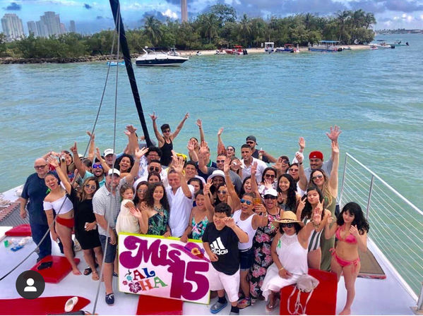 Large group celebrating a quinceañera on a white catamaran in turquoise bay, waving and smiling around a colorful "Mis 15" sign near a tree-lined island and distant skyline.