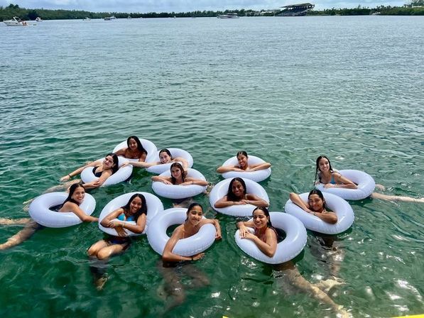 Group of friends floating on white inner tubes in clear green coastal water, smiling together near boats with a tree-lined shoreline in the distance — sunny tropical bay vibe.
