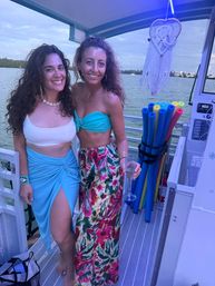 Two smiling women in tropical beachwear posing on a boat deck during an evening bay cruise, coastal water and sailboats in the distance, colorful pool noodles and a macrame dreamcatcher visible