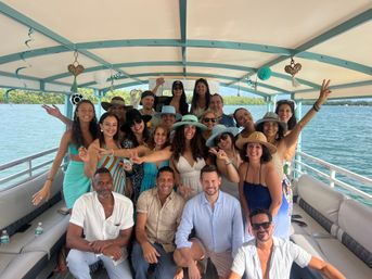 Large group of adults smiling and posing on a covered turquoise-and-white pontoon boat, many wearing sun hats and summer outfits, celebrating on calm blue water near a tree-lined shore.