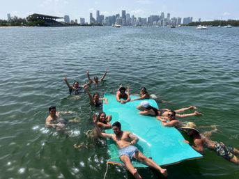 Group of friends swimming and lounging on a bright turquoise floating mat in Biscayne Bay with the Miami skyline and anchored yachts in the background on a sunny day.