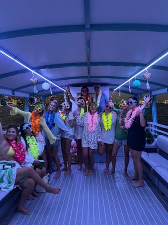 Nighttime pontoon boat party with a group wearing colorful leis and novelty sunglasses, posing barefoot under purple LED lights with city skyline and water in the background.