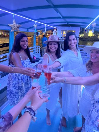 Friends toasting pink champagne on a covered night boat cruise, barefoot on deck under blue string lights with city waterfront in background