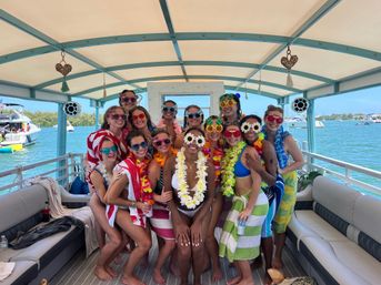 Group of friends in colorful swimsuits, leis and playful novelty sunglasses posing and smiling on a covered party boat over turquoise tropical bay with other boats in the background.