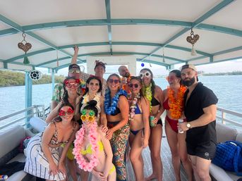 Group of friends in colorful swimsuits, leis and novelty sunglasses posing on a covered pontoon boat over calm blue water — fun summer boat party on a tropical waterway with shoreline in the distance.