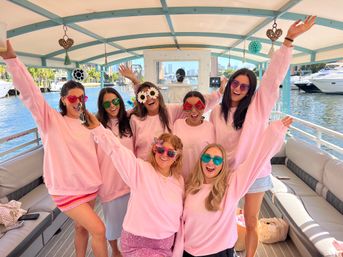 Seven friends in matching pink sweatshirts and playful sunglasses cheering with arms raised aboard a sunny pontoon boat at a marina, waterfront and city skyline visible in the background.