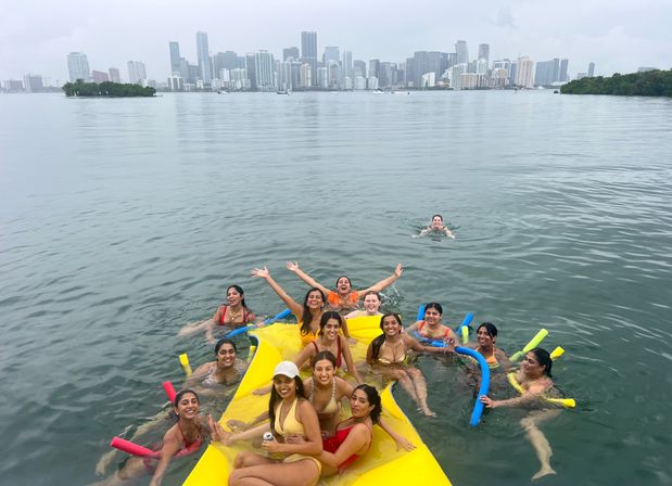 Smiling group of people in swimsuits lounging on a large yellow floating raft with colorful pool noodles in a calm bay, Miami skyline and overcast sky in the background.