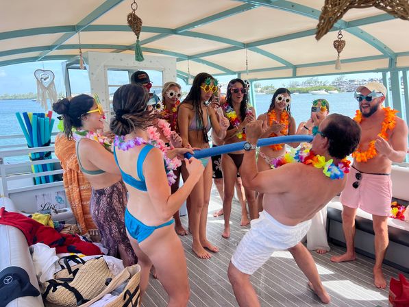 Adults in swimsuits and colorful leis playing limbo with a pool noodle on a covered party boat deck, wearing fun sunglasses and tropical decorations with turquoise ocean in the background.