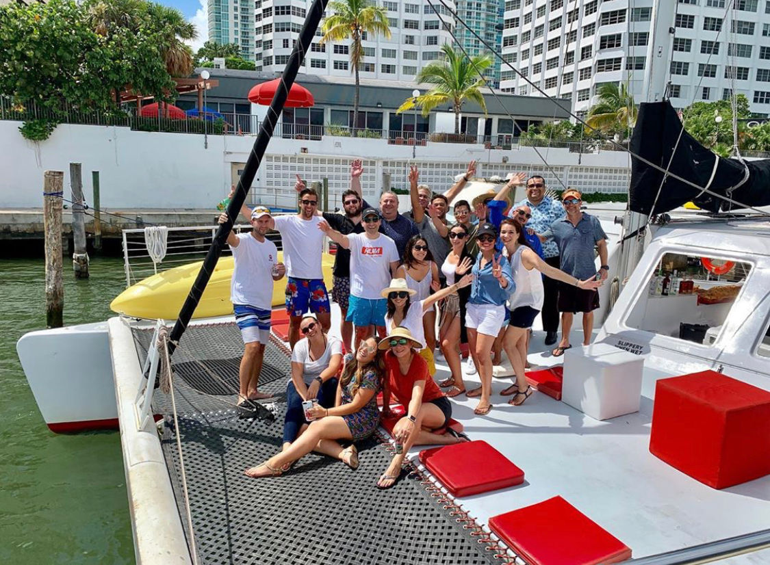 Happy group of adults waving and posing on a white catamaran at a sunny waterfront marina with palm trees and high-rise condos — tropical boat outing.